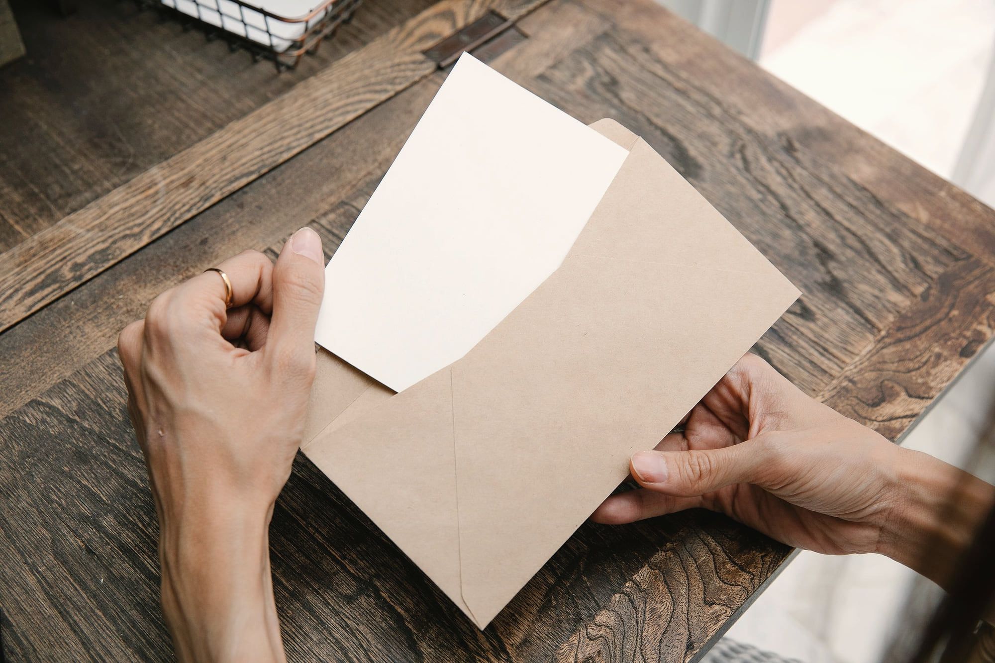 Deux mains sortent une carte d’une enveloppe kraft, sur une table en bois.