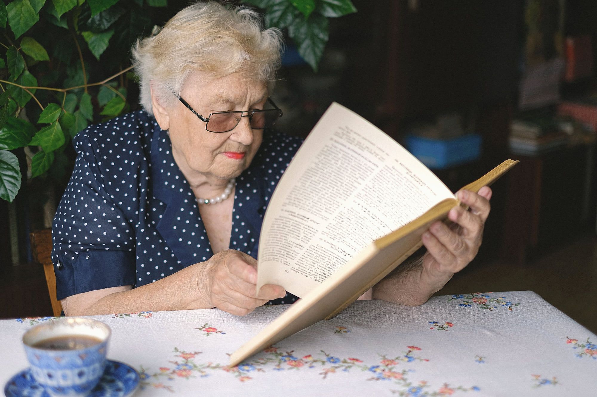 Une femme âgée lit un livre à la table du salon, près d’une tasse de thé.