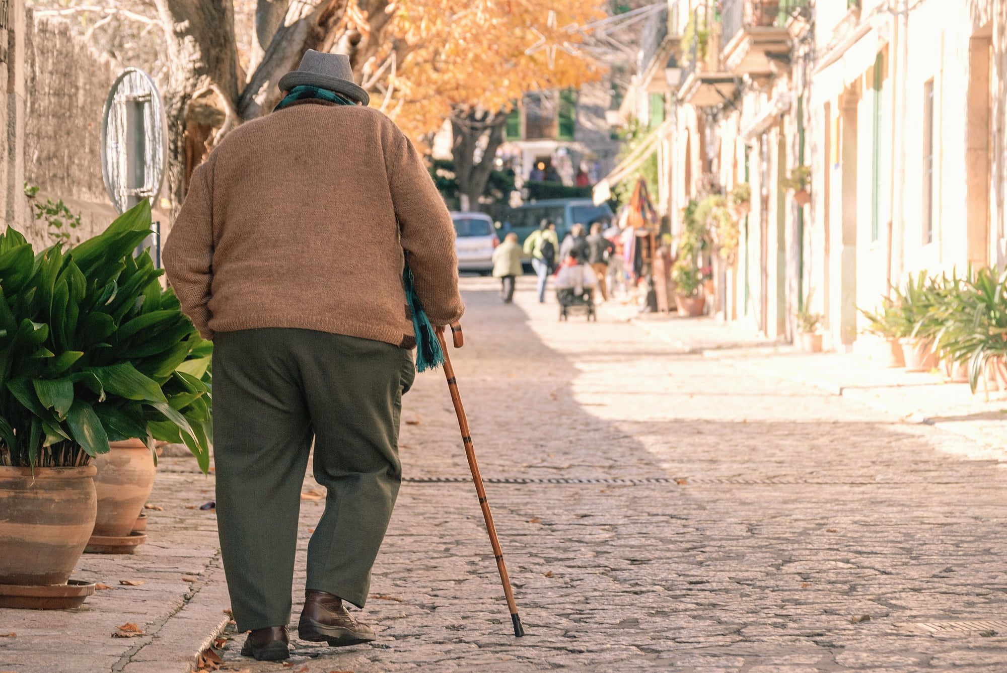 Un homme âgé marche avec une canne dans une rue pavée, en automne.